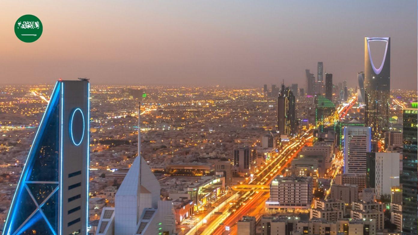 Rozświetlona panorama Rijadu o zmierzchu z Kingdom Centre, świecącymi autostradami i ikoną flagi Arabii Saudyjskiej.​​ 