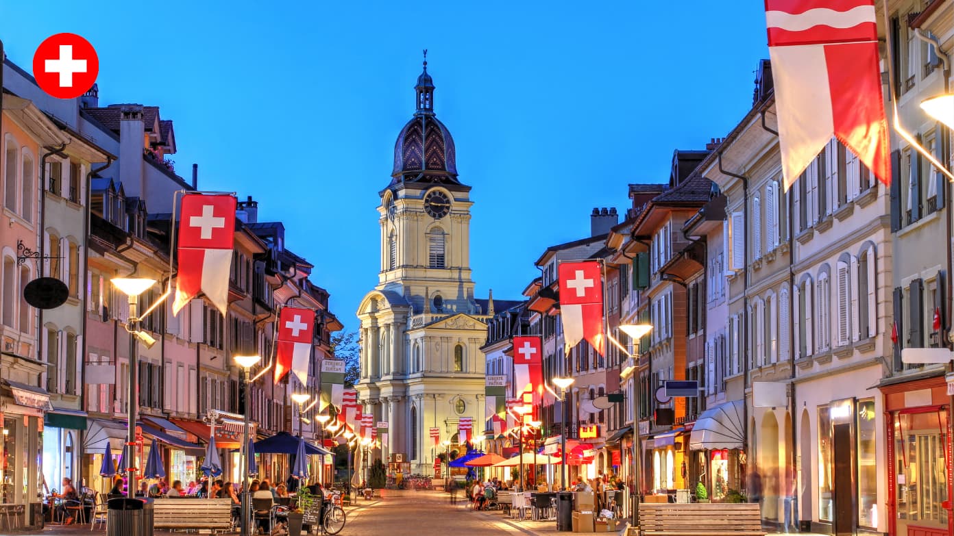 Charming street in Morges, Switzerland decorated with Swiss flags leading to a historic church tower at twilight​​ 