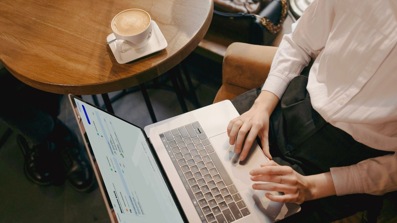 Top-down view of a professional working on a laptop next to a cup of coffee on a round wooden table.