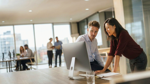 Twee collega's die samen werken op een desktopcomputer in een modern open kantoor.​​ 