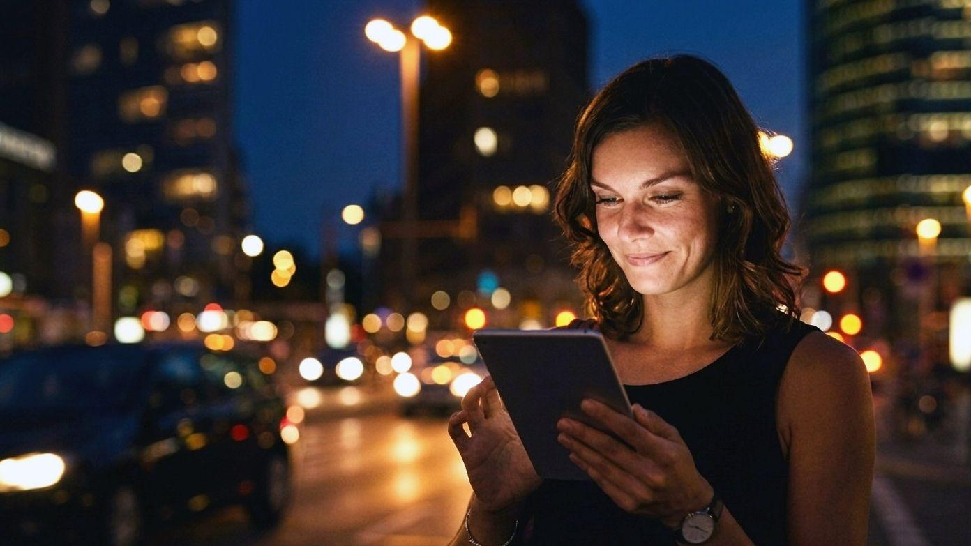 Glimlachende vrouw die 's nachts op een stadsstraat met gloeiende stadslichten een tablet gebruikt.​​ 