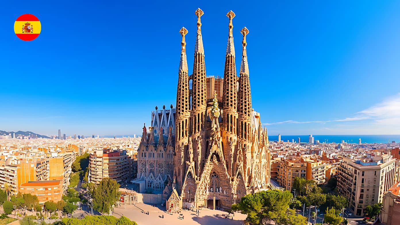 Aerial view of the towering Sagrada Família cathedral in Barcelona under a bright blue sky with a Spanish flag icon.​​ 